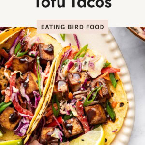 Close-up of two Asian-inspired tofu tacos topped with colorful slaw, scallions, sesame seeds, and lime wedges on a white plate.