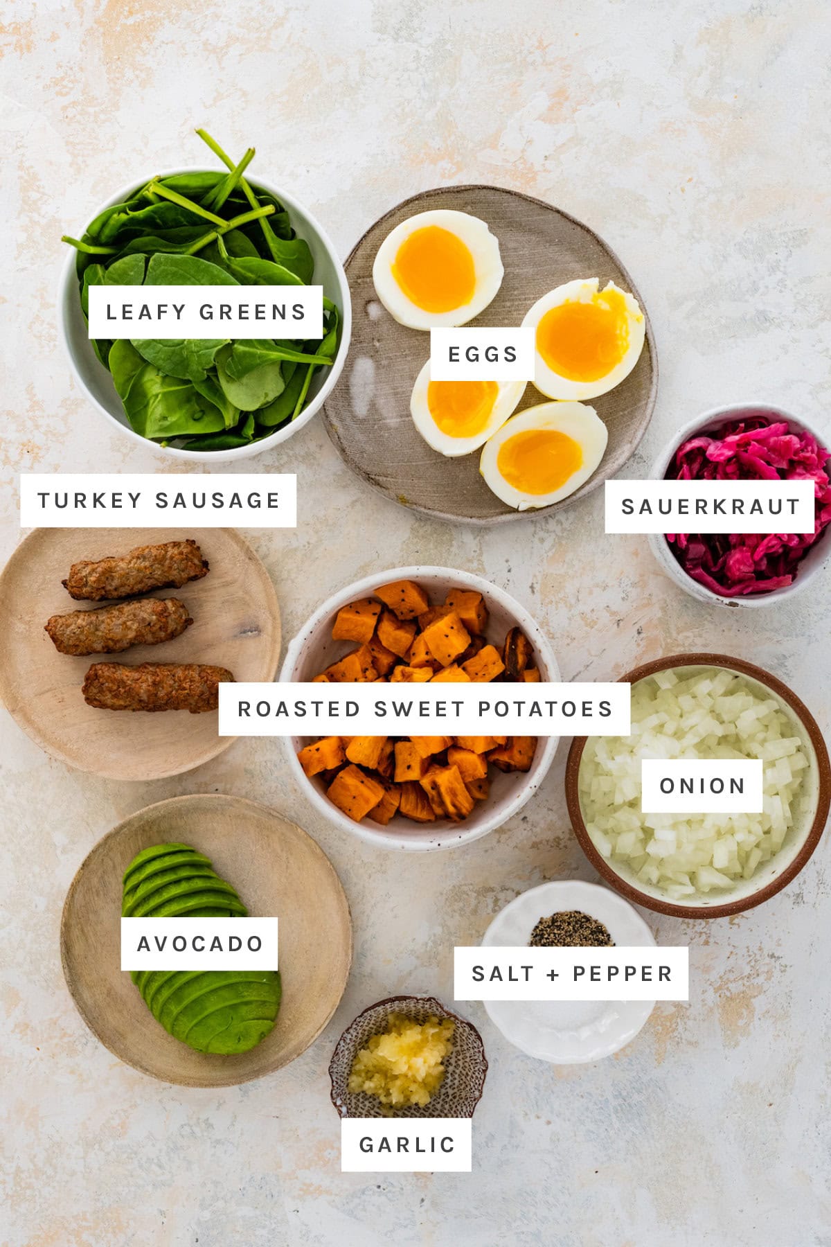 Ingredients measured out to make Breakfast Bowl: leafy greens, eggs, sauerkraut, turkey sausage, roasted sweet potatoes, onion, avocado, garlic, salt and pepper.