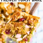 A slice of french toast bake on a wooden spoon with a casserole dish of the french toast back in the background. Bake is topped with cranberries and almond slices.
