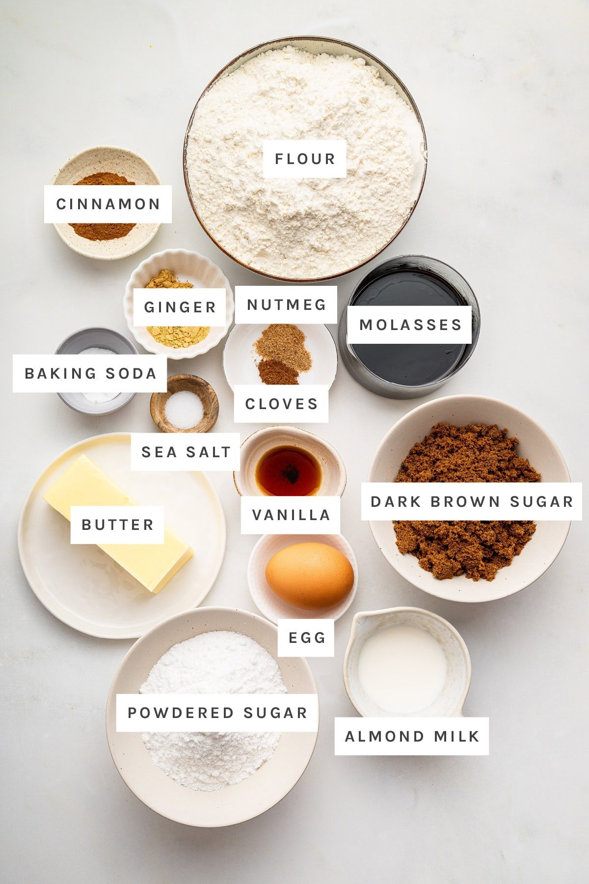 Ingredients measured out to make Gingerbread Cookies: cinnamon, flour, ginger, nutmeg, molasses, cloves, baking soda, sea salt, vanilla, dark brown sugar, butter, egg, powdered sugar and almond milk.