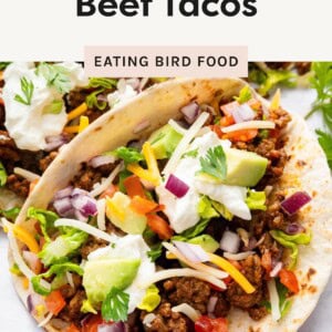 Close-up of a loaded soft taco with seasoned beef, shredded cheese, avocado, sour cream, diced tomatoes, lettuce, and red onion.