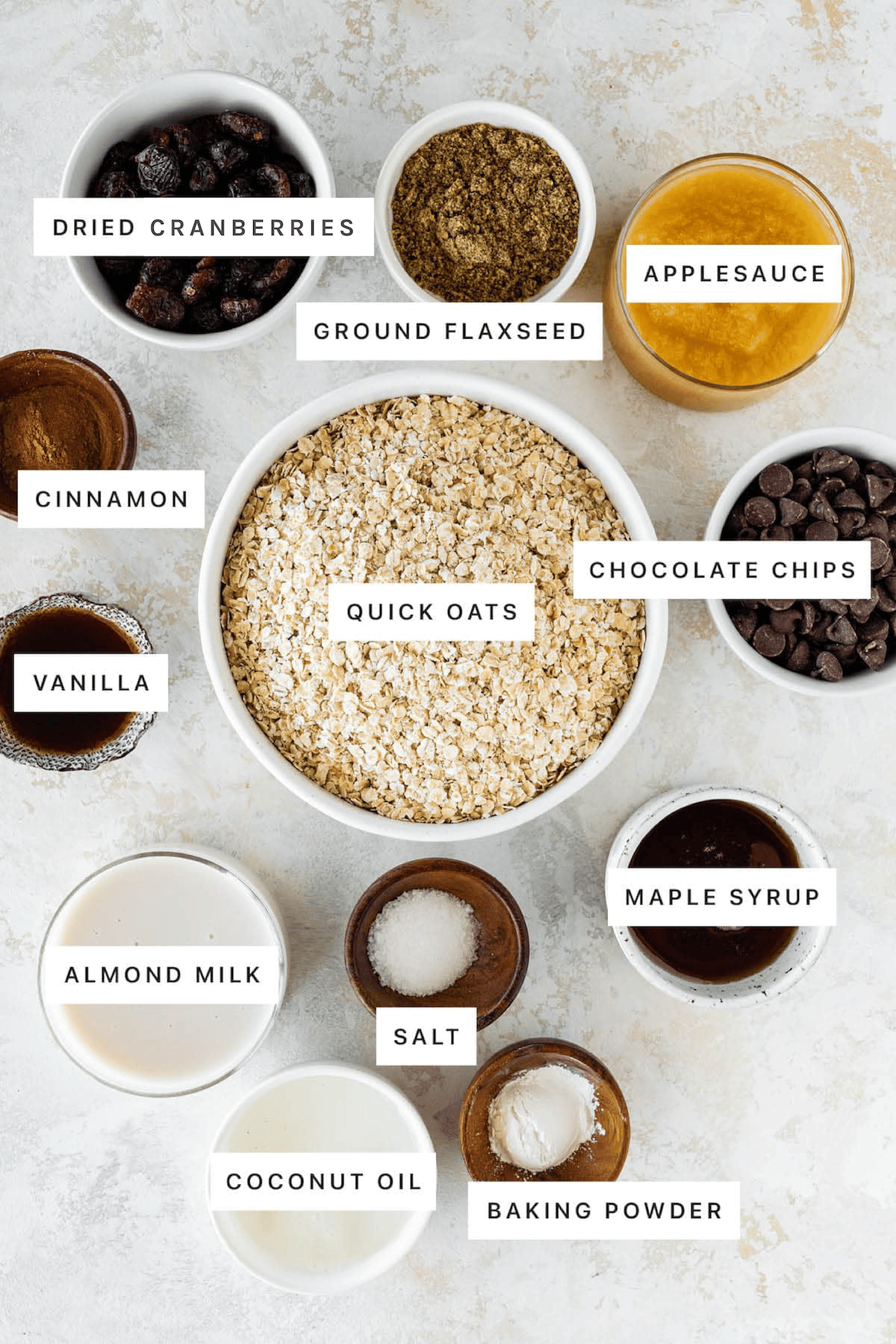 Ingredients measured out to make Oatmeal Breakfast Bars: dried cranberries, ground flaxseed, applesauce, cinnamon, quick oats, chocolate chips, vanilla, maple syrup, salt, almond milk, coconut oil and baking powder.