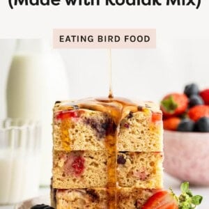 A tall stack of berry-filled sheet pan pancakes drizzled with syrup, served on a plate with fresh strawberries and blueberries.