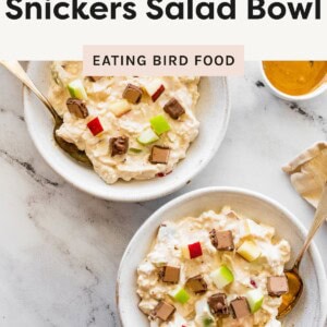 Two bowls of high protein Snickers salad topped with chopped apples and chocolate pieces on a marble surface with spoons and peanut butter on the side.