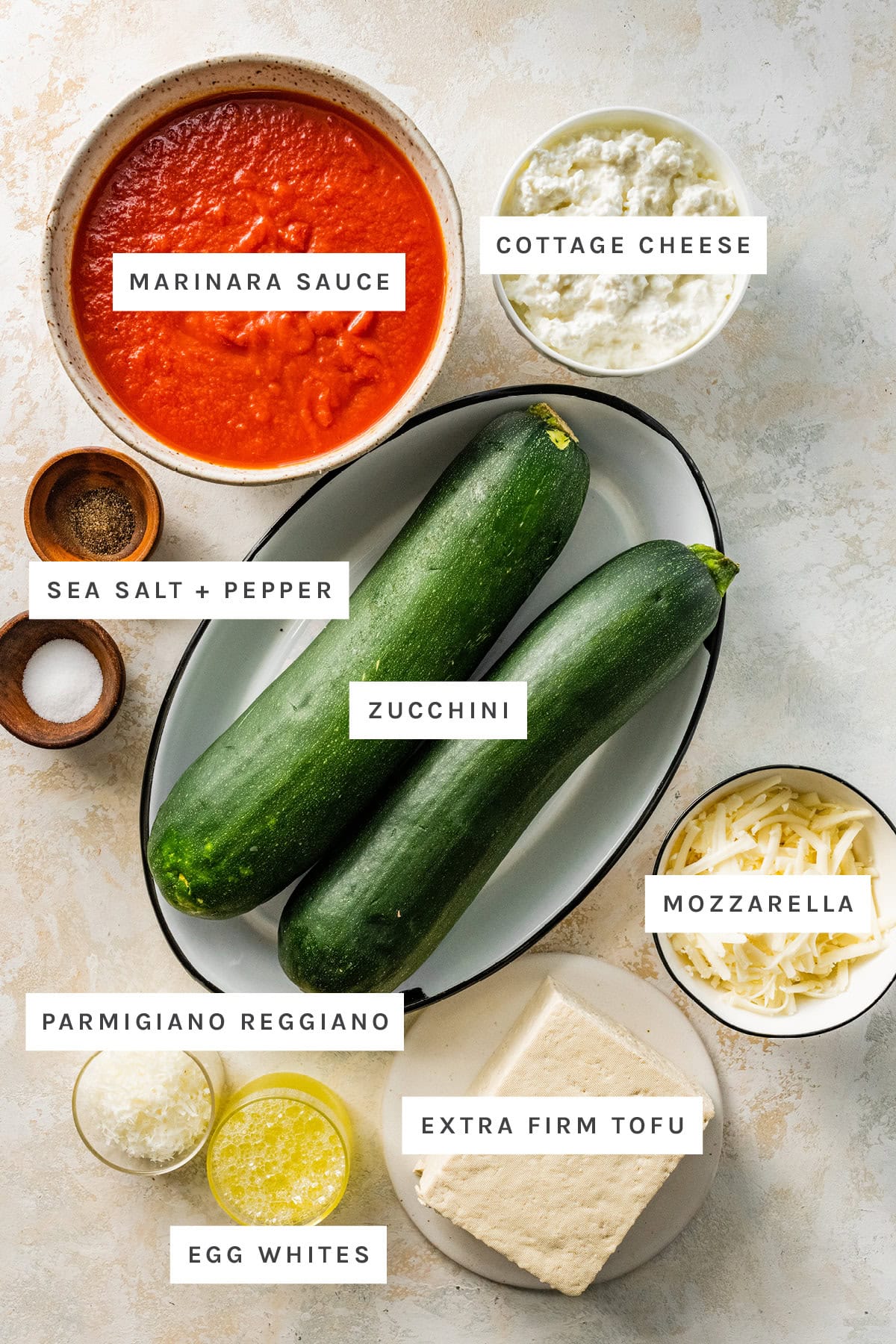 Ingredients measured out to make Tofu Lasagna with Zucchini Noodles: marinara sauce, cottage cheese, sea salt, pepper, zucchini, parmigiano reggiano, extra firm tofu, egg whites and mozzarella.