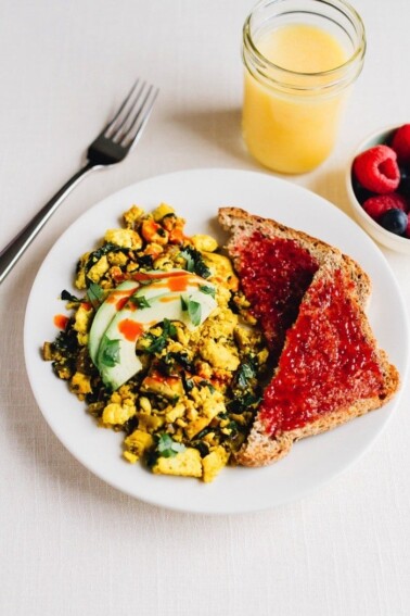 Turmeric tofu scramble on a plate with avocado on top. Two slices of bread with jam on the side. A bowl of raspberries and blueberries is next to the plate. Glass of orange juice and fork also on the side.