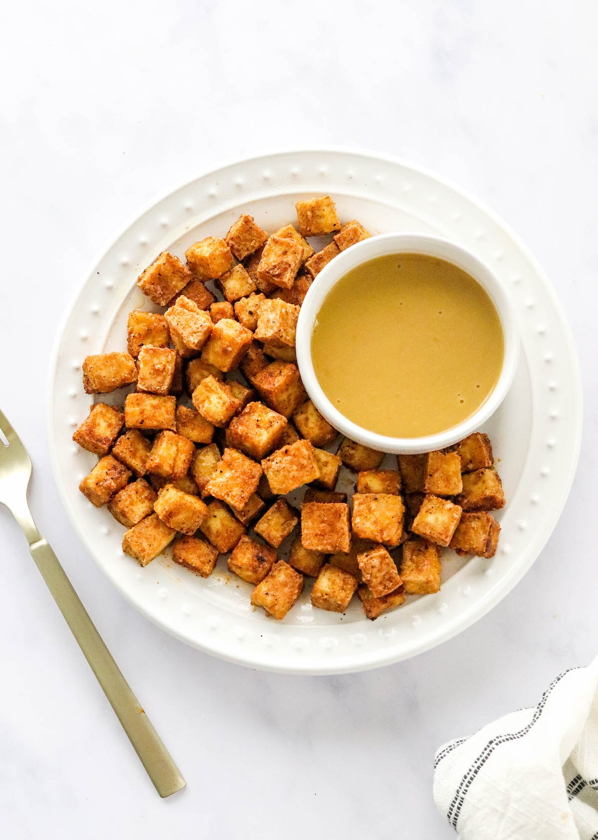 An overhead photo of a plate of air fried tofu with a ramekin of dipping sauce. A gold fork is next to the plate.