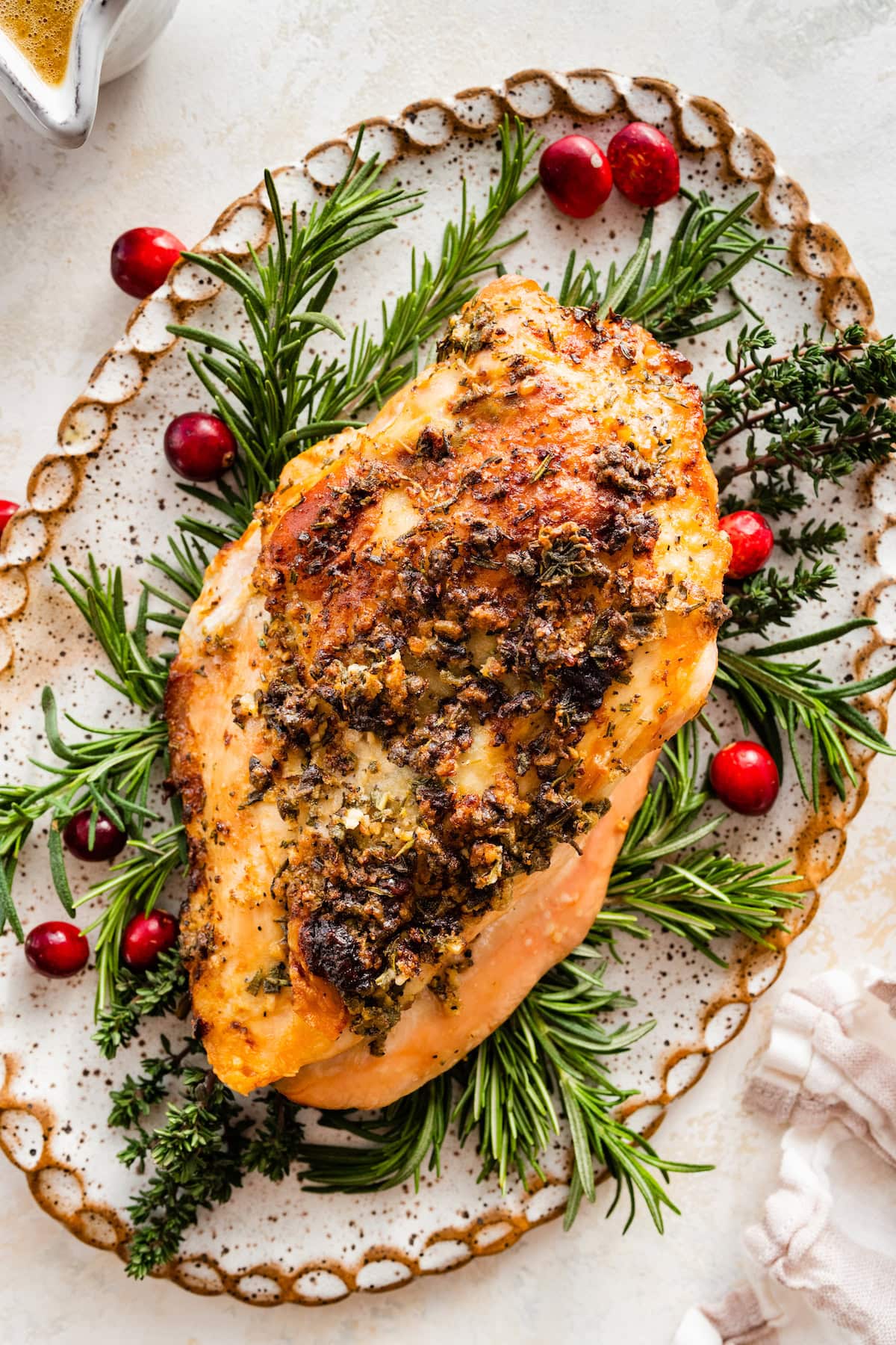 An un-sliced air fryer turkey breast sitting on a bed of fresh herbs on a white platter. Fresh cranberries are sprinkled around the platter.
