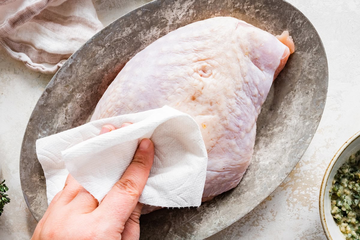 A woman's hand patting a boneless turkey breast dry with a paper towel. The turkey breast is sitting on a silver platter.
