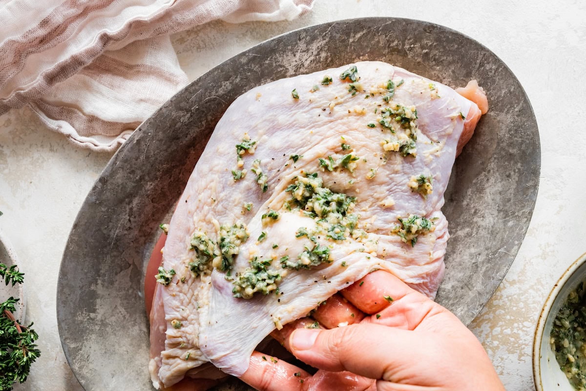 A woman's hand seasoning a turkey breast with butter, garlic and herbs under the skin. The turkey breast is sitting on an silver platter.