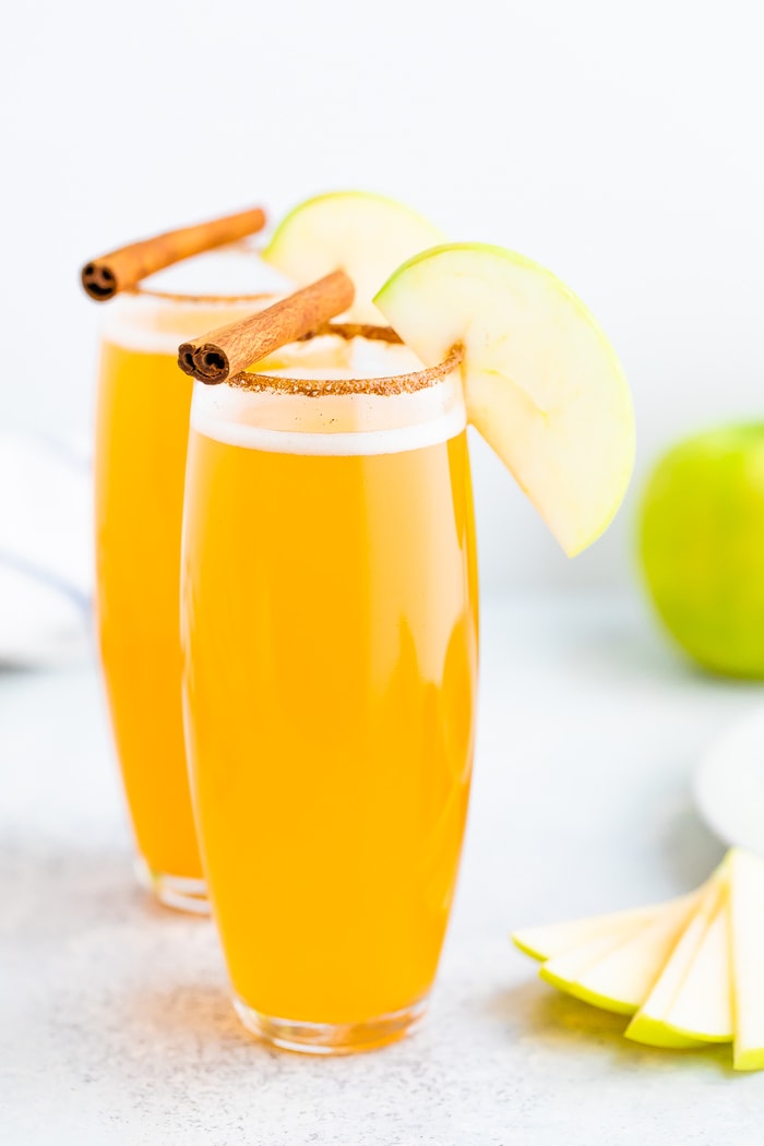 Two tall glasses of apple cider mimosas with a cinnamon sugar rim, cinnamon sticks and an apple slice for garnish.