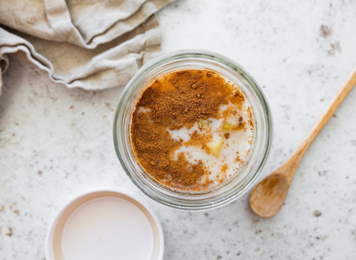 Overhead view of a glass jar with diced apples, milk, and cinnamon sprinkled on top before mixing.