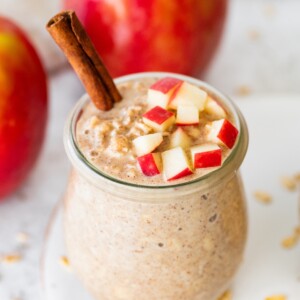 Two jars of apple cinnamon overnight oats on a white plate topped with chopped apples and cinnamon sticks with oats scattered around.