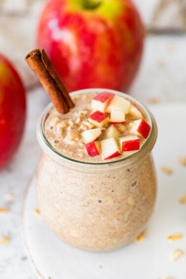 Two jars of apple cinnamon overnight oats on a white plate topped with chopped apples and cinnamon sticks with oats scattered around.