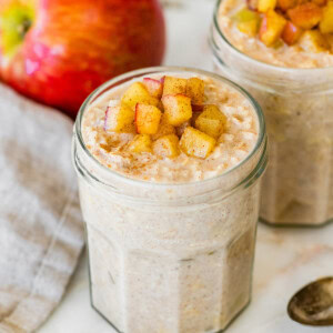 Two jars of apple cinnamon overnight oats topped with diced apples, with a fresh apple behind the jars.