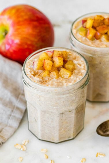 Two jars of apple cinnamon overnight oats topped with diced apples, with a fresh apple behind the jars.
