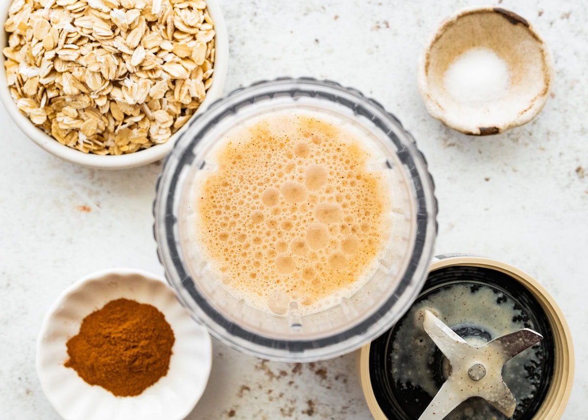Blender cup filled with the almond milk, almond butter and apple juice mixture for apple cinnamon overnight oats surrounded by oats and cinnamon.
