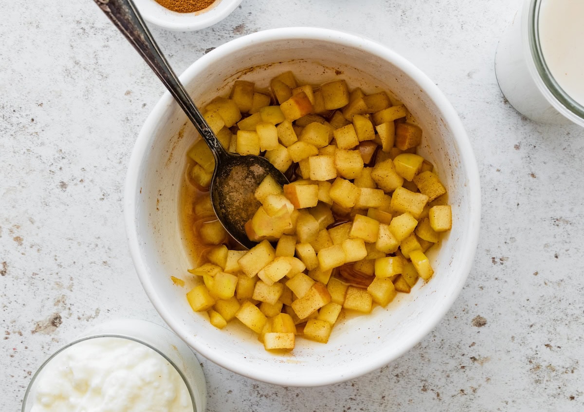 White bowl filled with diced apples cooked with cinnamon and liquid, with a spoon inside.