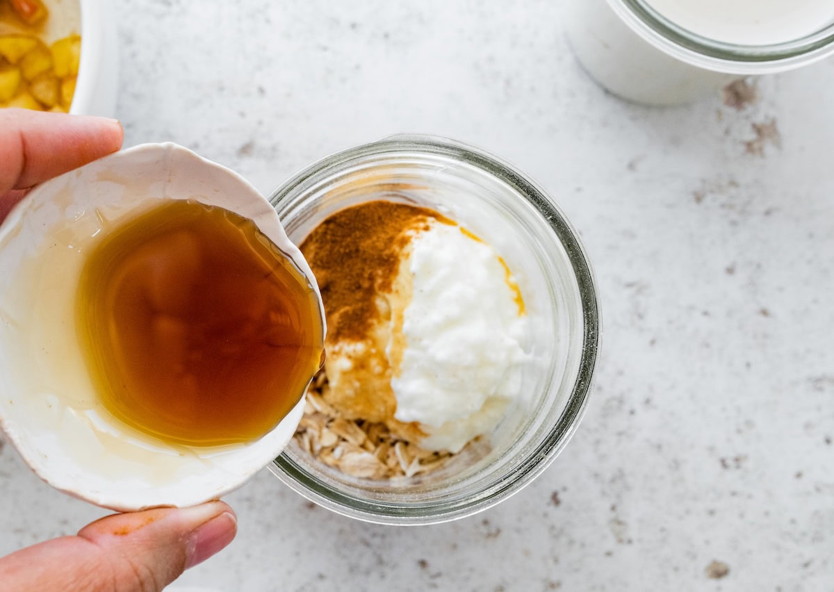 Hand pouring maple syrup into a glass jar containing oats, yogurt, and cinnamon.