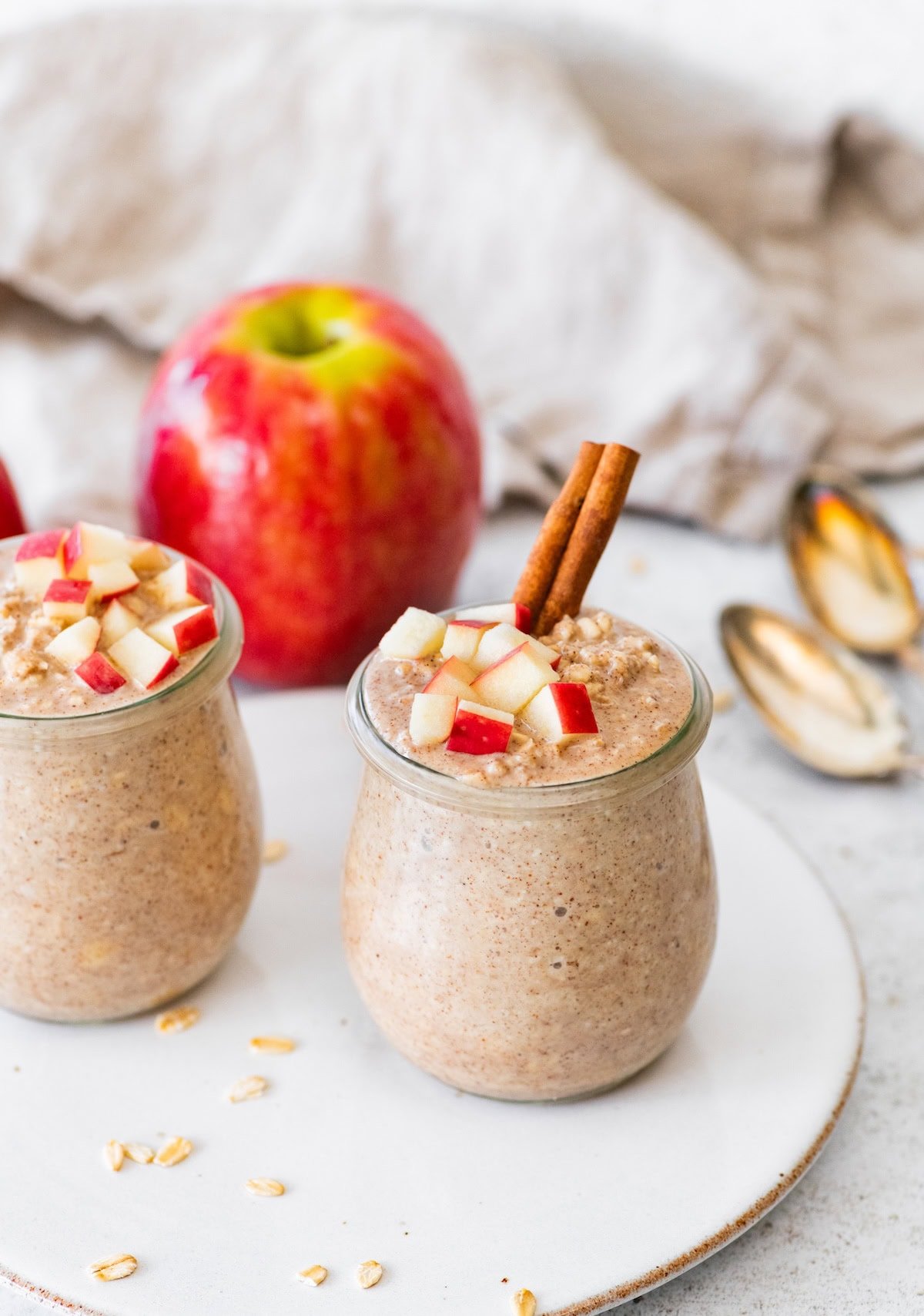 Two jars of apple cinnamon overnight oats topped with chopped apples and cinnamon sticks with a fresh apple in the background.