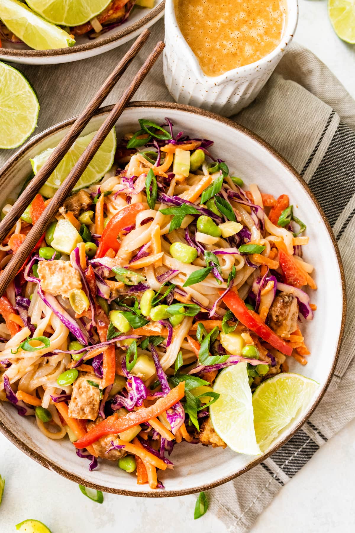 All ingredients for the asian noodle bowl mixed together in a white bowl with wooden chopsticks and lime wedges on the side.