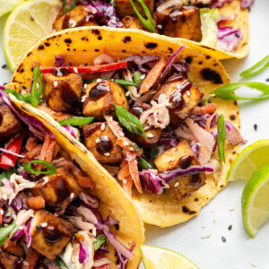 Three tofu tacos in yellow corn tortillas, loaded with slaw, tofu cubes, scallions, and sesame seeds, with lime slices in the background.