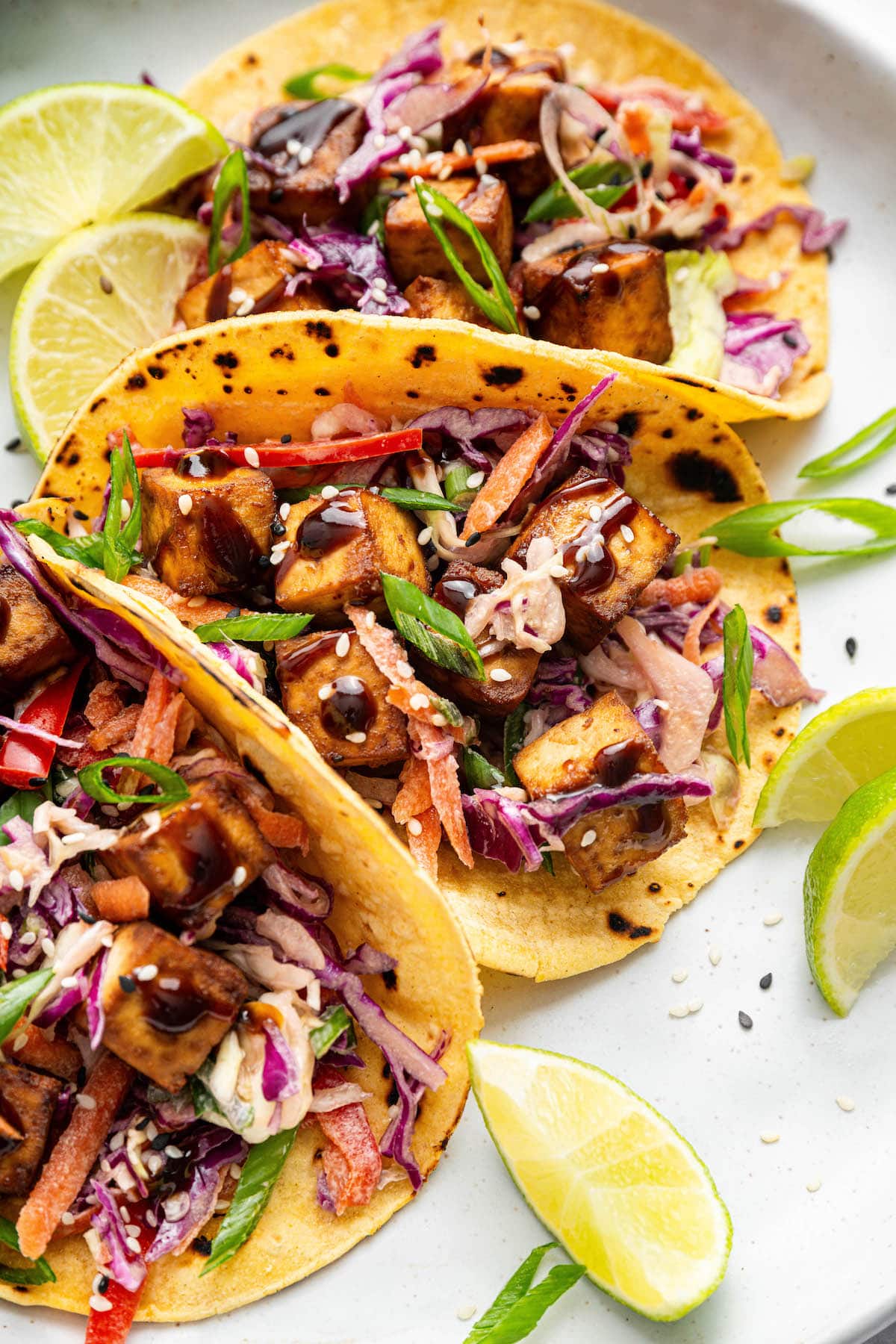 Three tofu tacos in yellow corn tortillas, loaded with slaw, tofu cubes, scallions, and sesame seeds, with lime slices in the background.