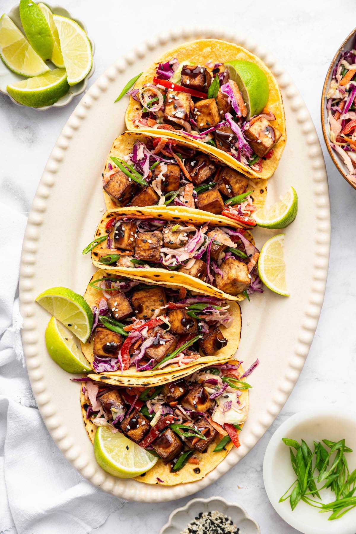 Six corn tofu tacos on an oval platter, filled with crispy tofu, crunchy Asian slaw, and garnished with lime wedges.