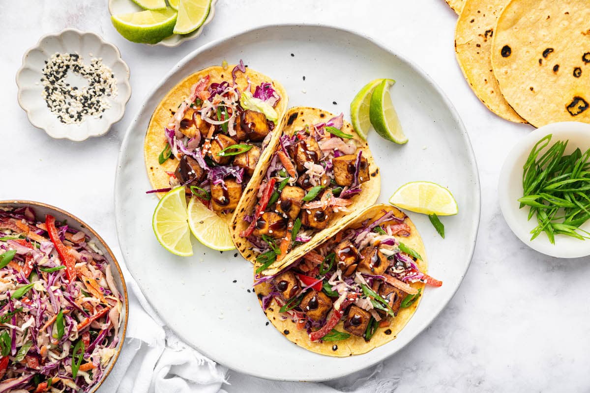 Three tofu tacos plated on a white dish, topped with slaw and garnished with lime wedges.