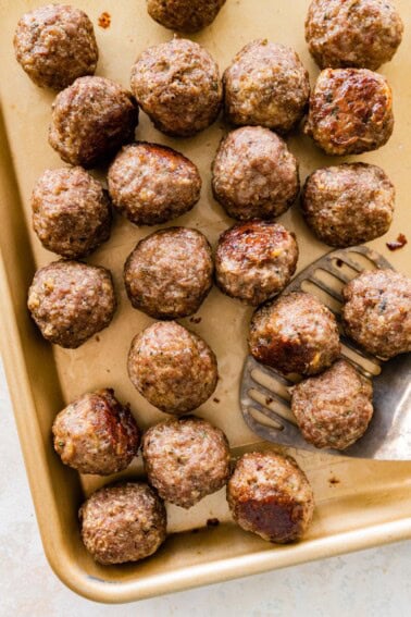 Baked meatballs resting on a parchment-lined sheet pan with browned tops and a metal spatula taking a few from the pan.