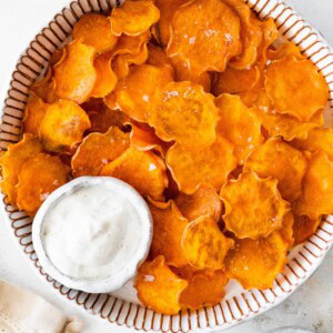 Homemade baked sweet potato chips served in a striped bowl with a side of dip.