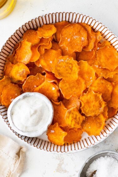 Homemade baked sweet potato chips served in a striped bowl with a side of dip.