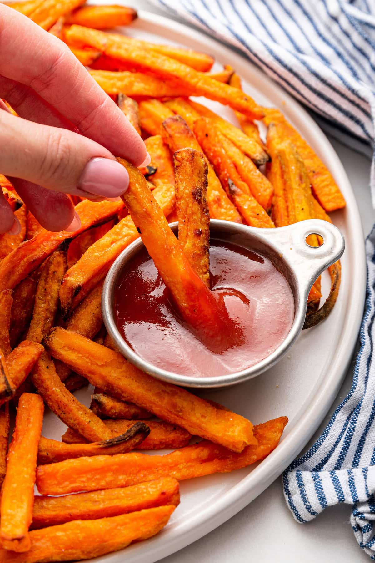 A womans hand dips a sweet potato fry in ketchup.