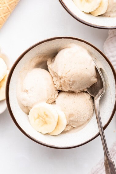 Three scoops of banana ice cream in a bowl with a metal spoon and two slices of banana.