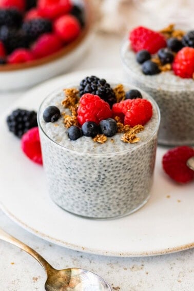 Glass of basil seed pudding topped with berries and granola on a white plate.