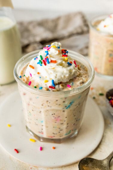A glass jar filled with birthday cake overnight oats, topped with whipped cream and colorful sprinkles, with a bottle of milk and another jar of oats in the background.