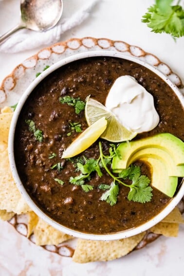 A bowl of black bean soup topped with avocado, cilantro, lime and vegan sour cream served alongside tortilla chips.