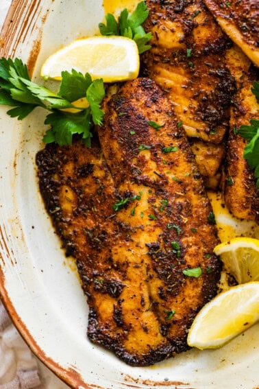 Blackened tilapia filets on a plate topped with fresh parsley and served with lemon wedges.