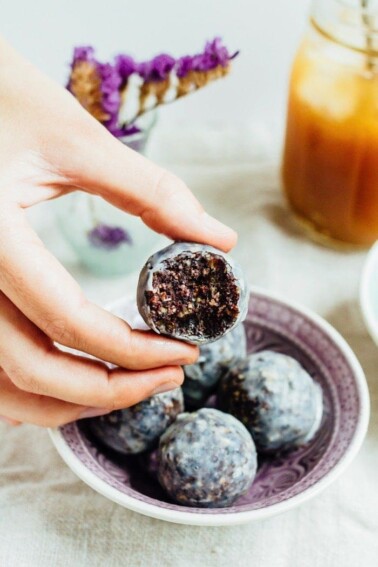 A hand holding a blueberry muffin donut hole with a bite taken out of it.
