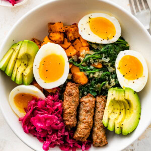 A breakfast bowl with 2 soft boiled eggs, 3 turkey sausage, sliced avocado, sauerkraut, roasted sweet potatoes and sautéed kale all in a white bowl.