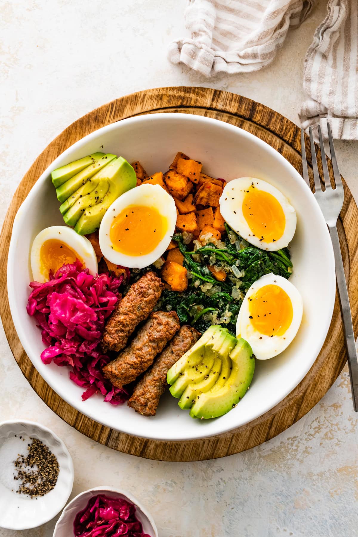 A breakfast bowl with 2 soft boiled eggs, 3 turkey sausage, sliced avocado, sauerkraut, roasted sweet potatoes and sautéed kale all in a white bowl.