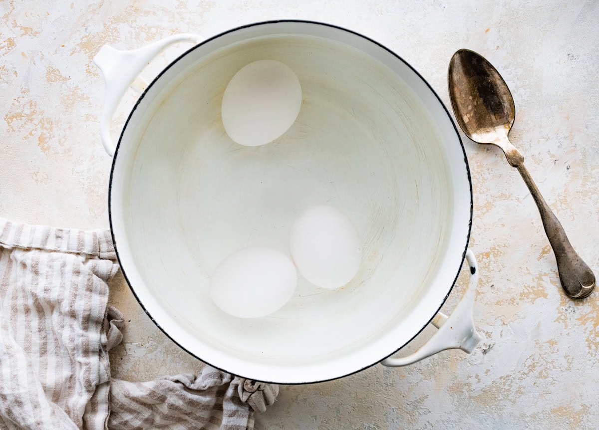 3 eggs cooking in a pot of water. There is a silver spoon next to the white pot.