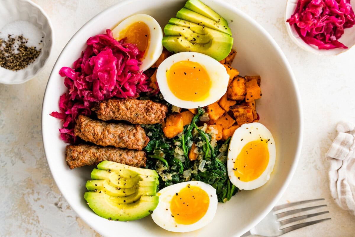 A breakfast bowl with 2 soft boiled eggs, 3 turkey sausage, sliced avocado, sauerkraut, roasted sweet potatoes and sautéed kale all in a white bowl.