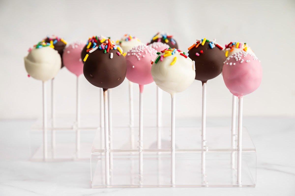 Chocolate and pink cake pops with colorful sprinkles arranged upright in a clear cake pop stand.