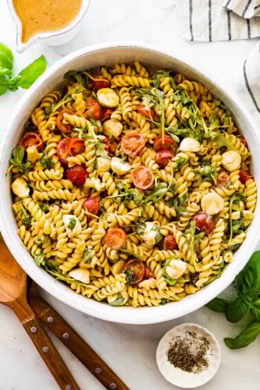 A serving bowl filled with caprese pasta salad. There are serving spoons next to the bowl.