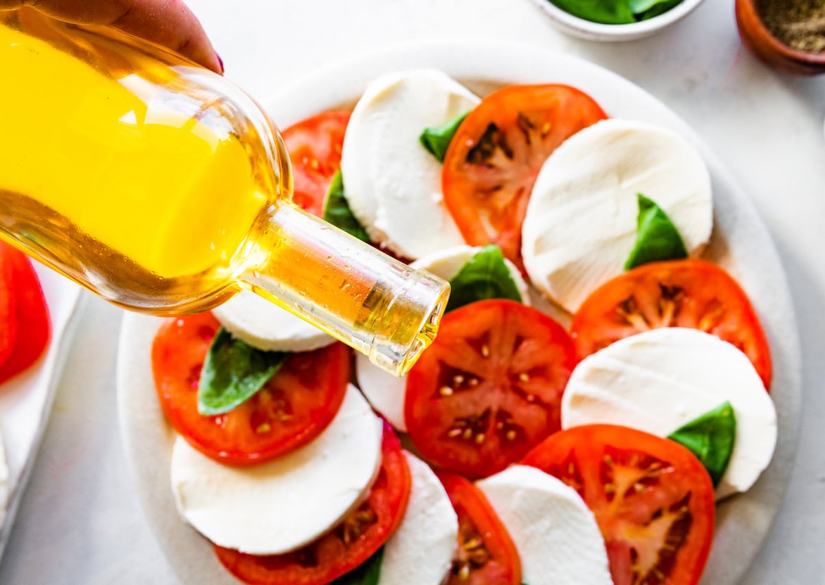 Olive oil is poured onto a large plate of a caprese salad with fresh tomatoes and mozzarella cheese.