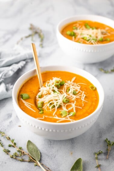 Two bowls of carrot parsnip soup garnished with scallions and shredded gruyere cheese.