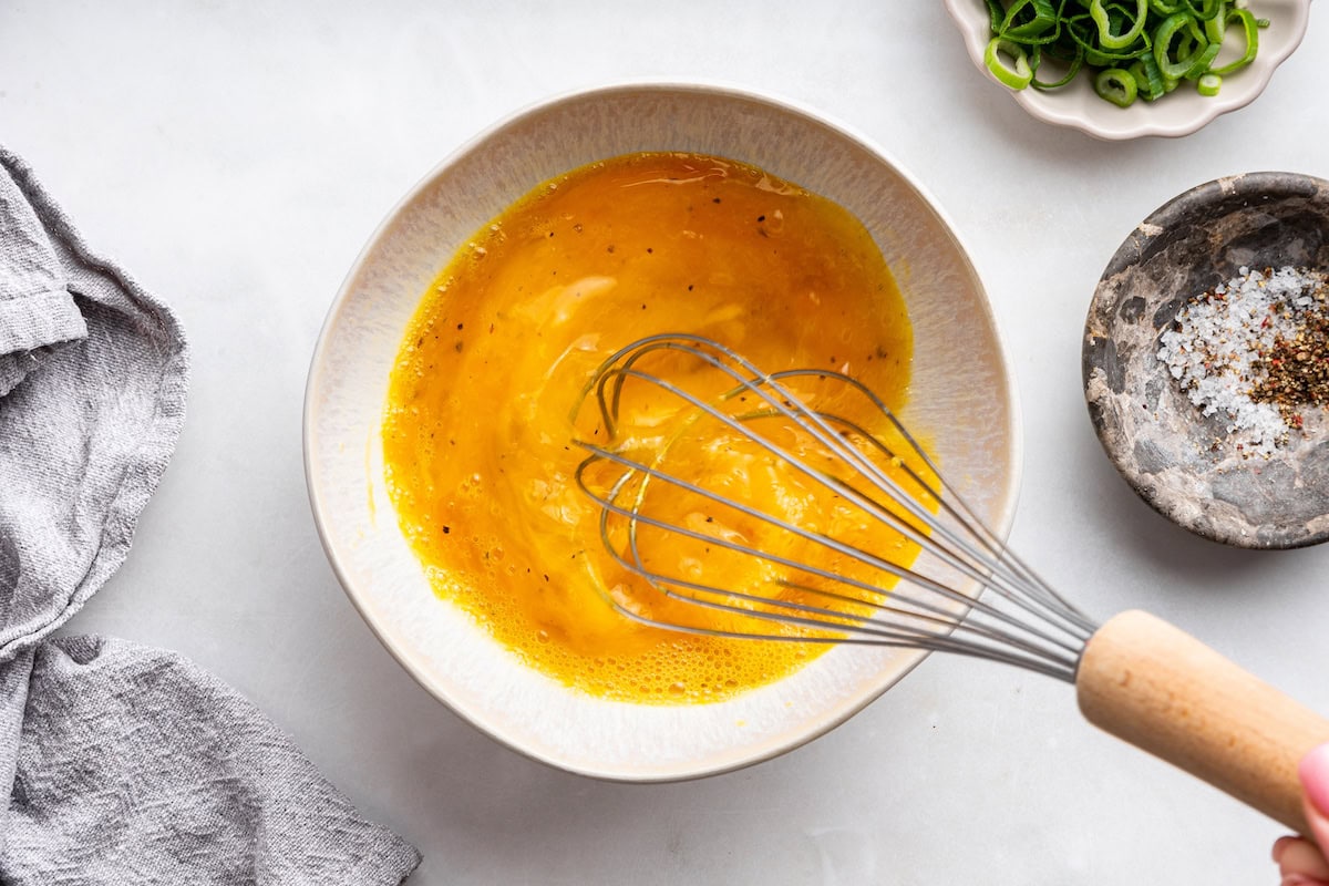 A metal whisk in a bowl whisking eggs.