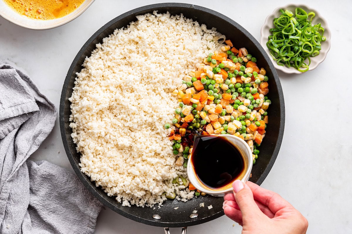 A woman's hand pours soy sauce into a skillet of cauliflower rice and a mix of frozen vegetables.