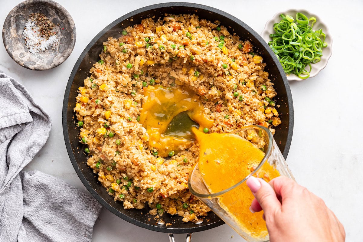 A woman's hand pours eggs into a skillet with cauliflower rice and mixed vegetables.